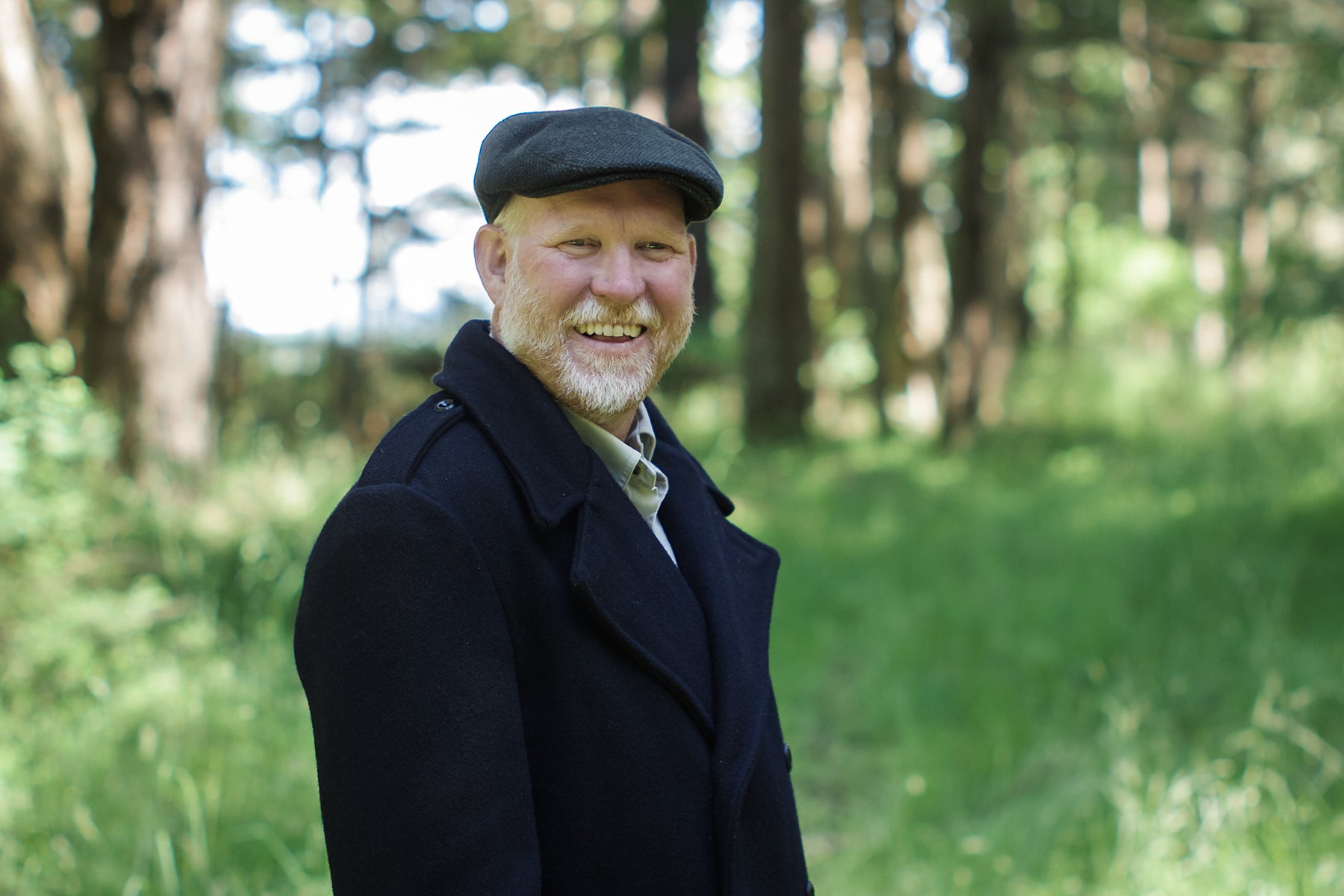 Photograph of a man in his late forties wearing a wool Thread Theory peacoat and a flat cap, smiling and looking at the camera. He's standing in front of trees and grass that are out of focus in the background. 