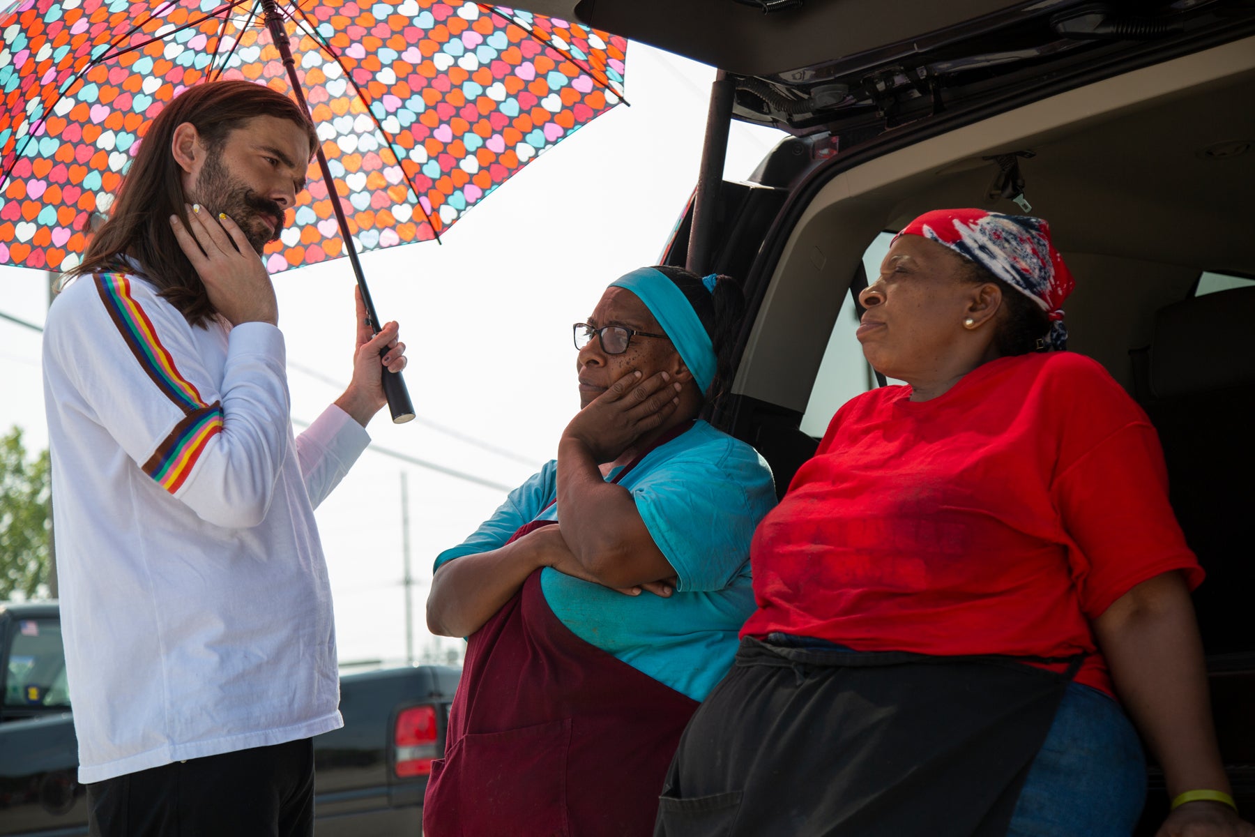 Jonathan holds an umbrella decorated with small hearts and speaks to two women who are sitting on the bumper of a truck in an episode of Queer Eye. 