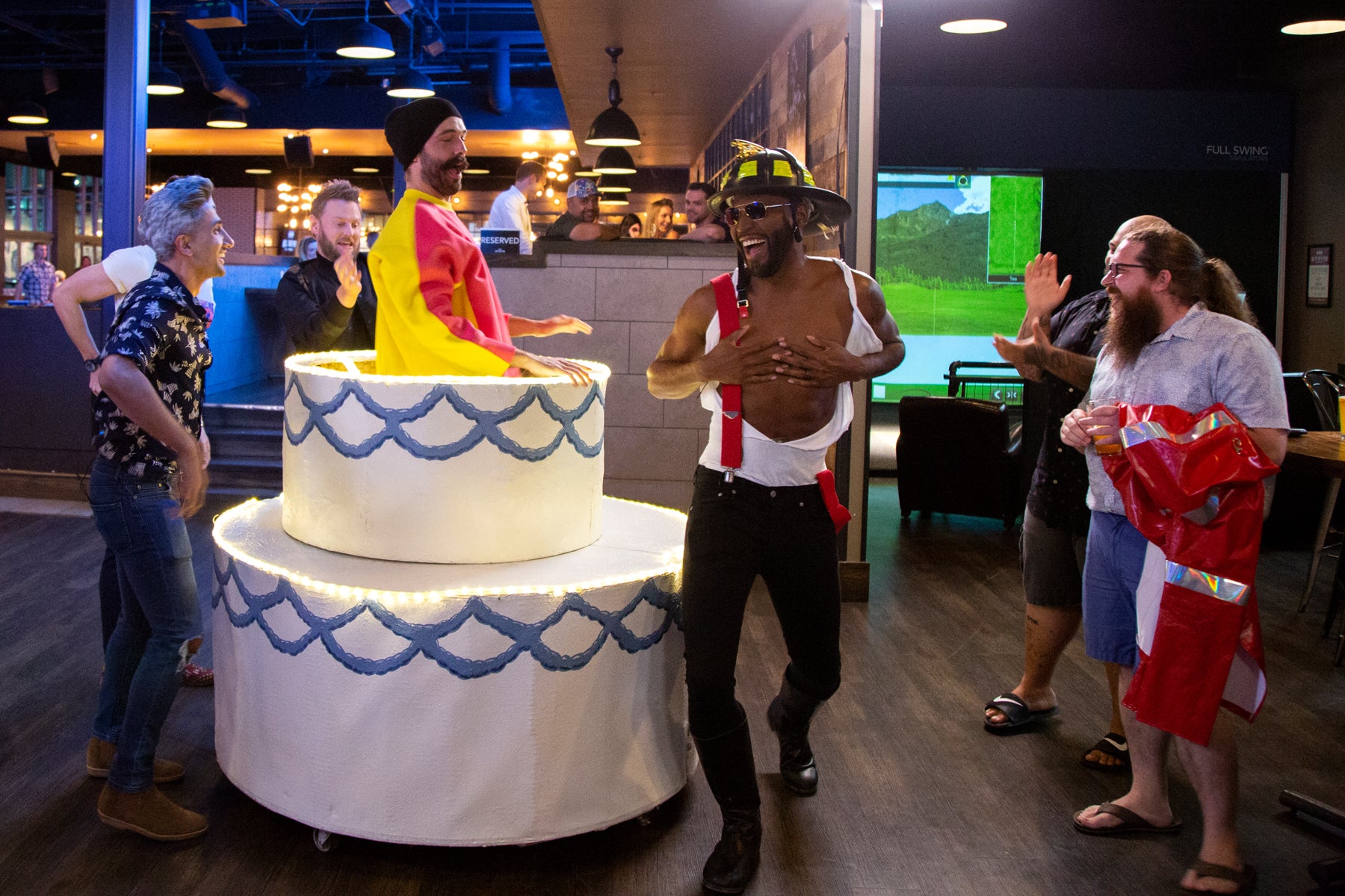 Jonathan stands in an oversized cake, and Karamo dances in a firefighter hat, while the cast of Queer Eye watches.