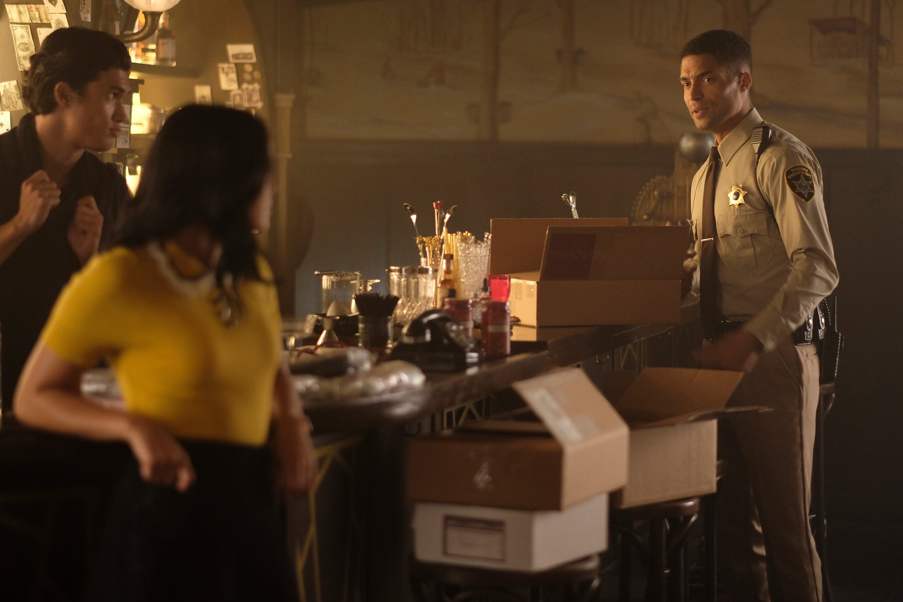 In the speakeasy, Reggie Mantle and Veronica Lodge look on as Sheriff Minetta looks through boxes on the bar.