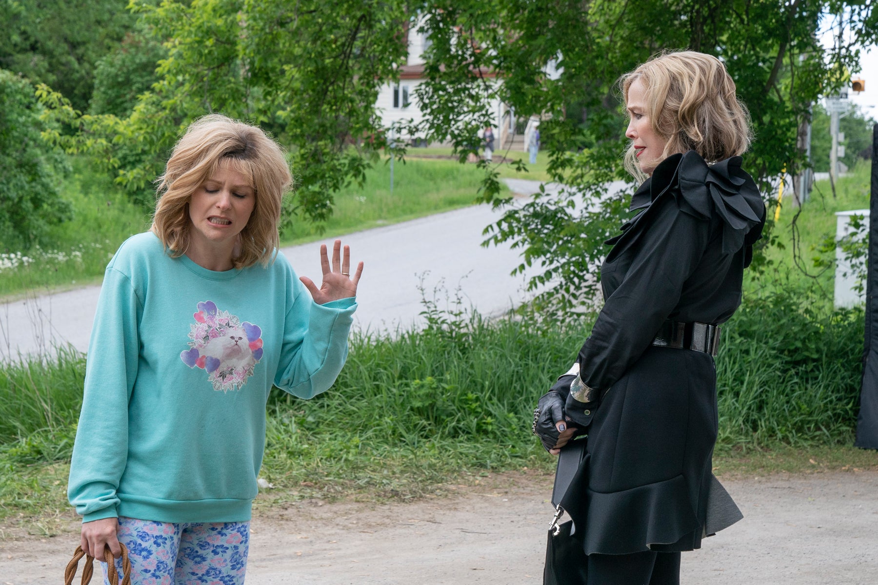 Jocelyn Schitt looks distressed while holding up a hand to Moira Rose as both women talk outside next to a road.