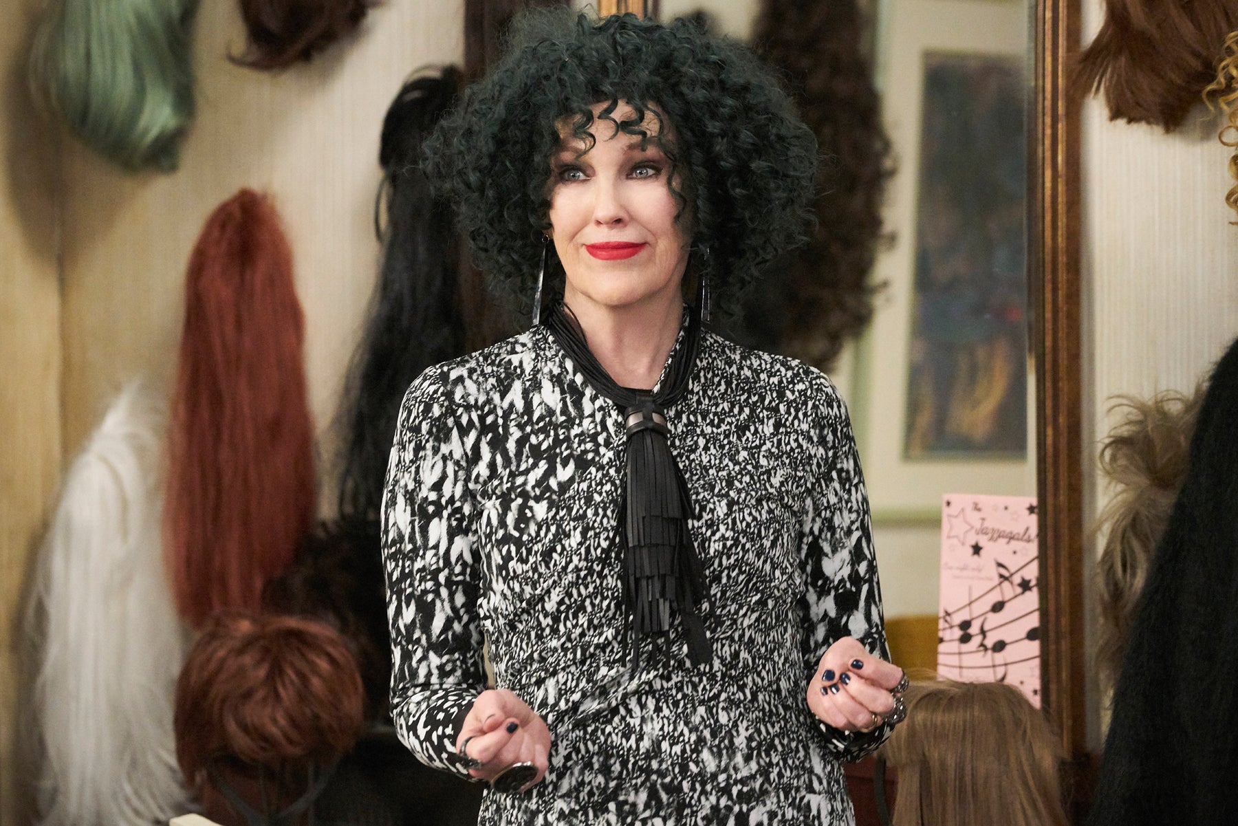 Moira Rose wears a curly black wig and stands in front of her “wig wall” in her motel room.