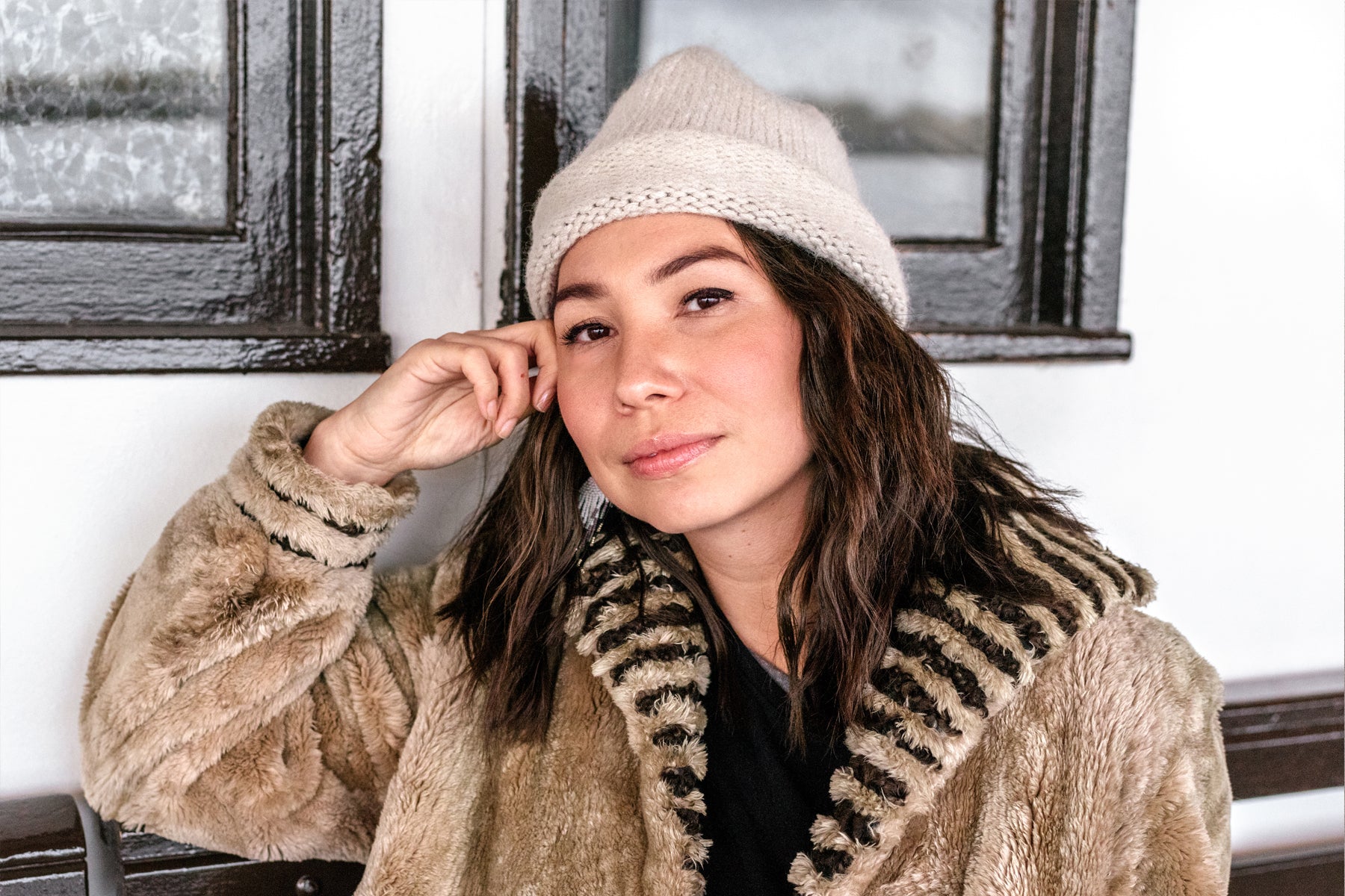 Portrait of designer Sage Paul sitting down in a fur coat and wool toque resting her head on her hand.