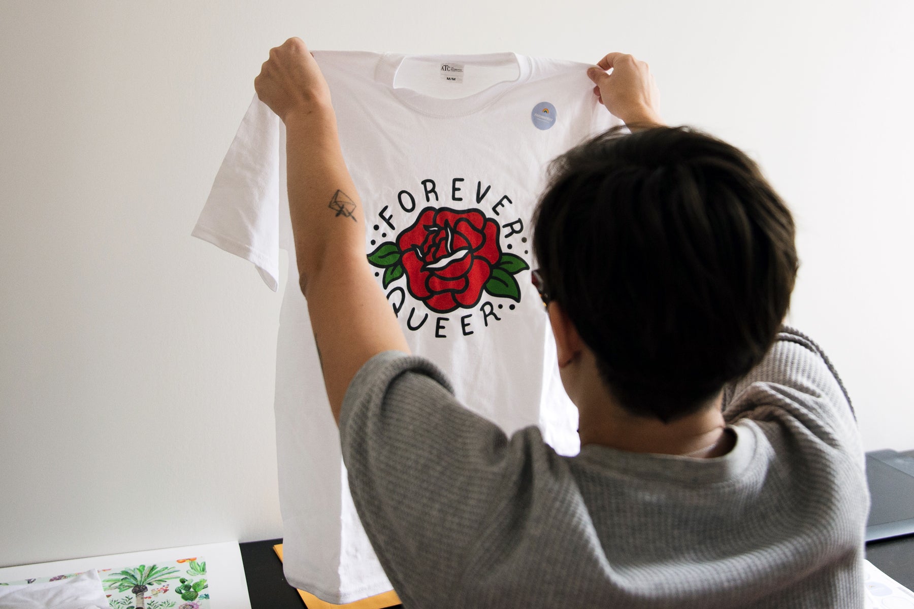 Image of designer Liz Bertorelli holding up one of her custom t shirts that reads Forever Queer.