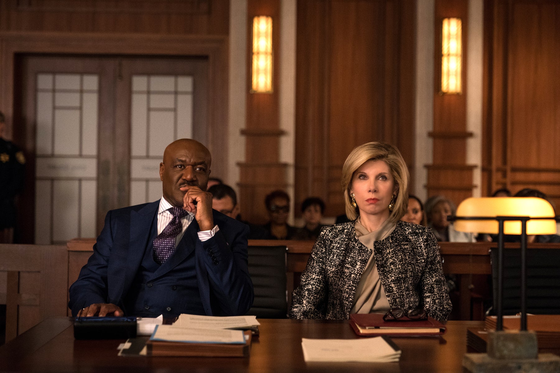 Adrian and Diane sit facing forward at a wooden table in court. They both look serious.