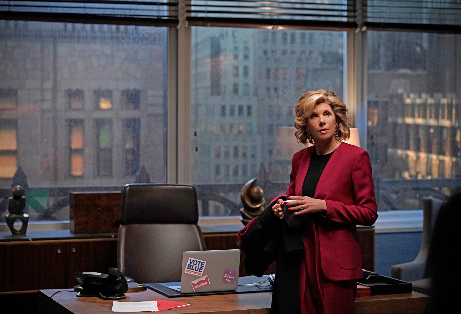 Diane stands in her office, facing away from the windows that show it is raining and gloomy outside. On her desk, there is a laptop with multiple stickers that say things like “Vote Blue” and “MeToo.”