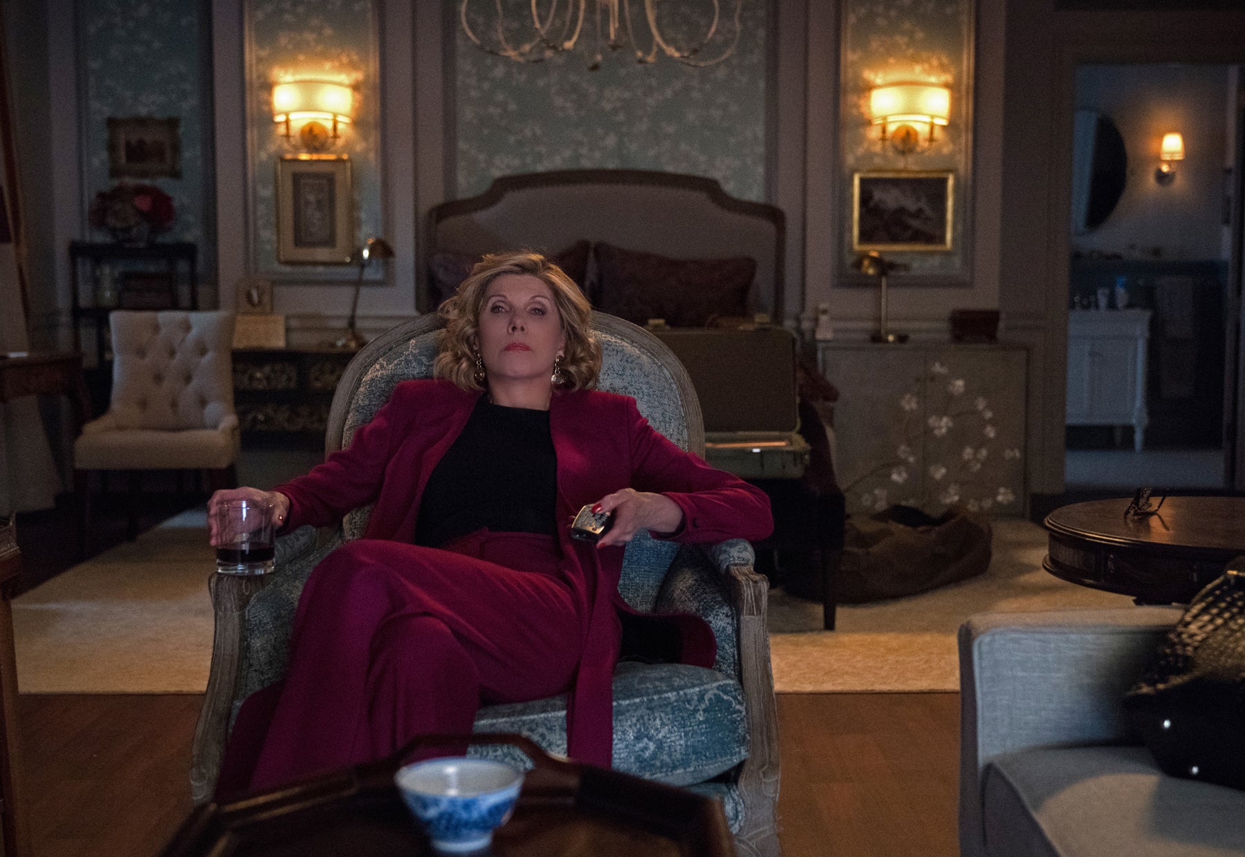 Diane sits slumped in a chair in her bedroom with a glass of liquor in one hand and the remote in the other. She looks tired, as if she has just had a long day.