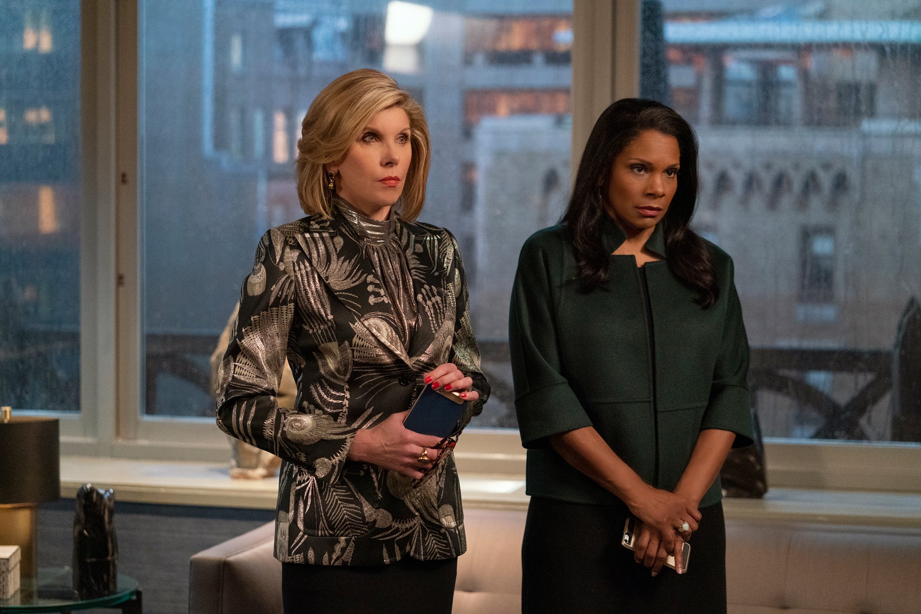 Diane Lockhart and Liz Reddick look serious during a meeting. Diane is wearing a shiny, patterned blazer and shirt with black pants. Liz is wearing a voluminous dark green coat and black pants. They are both holding their cellphones.