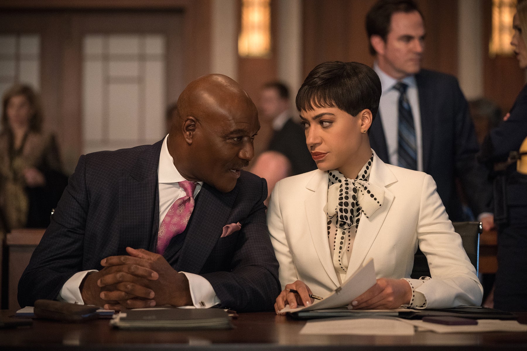 Adrian Boseman leans over to talk to Lucca Quinn in court during a trial where they are defending Chum Hum. They both look annoyed because in the episode, their clients are being difficult. 