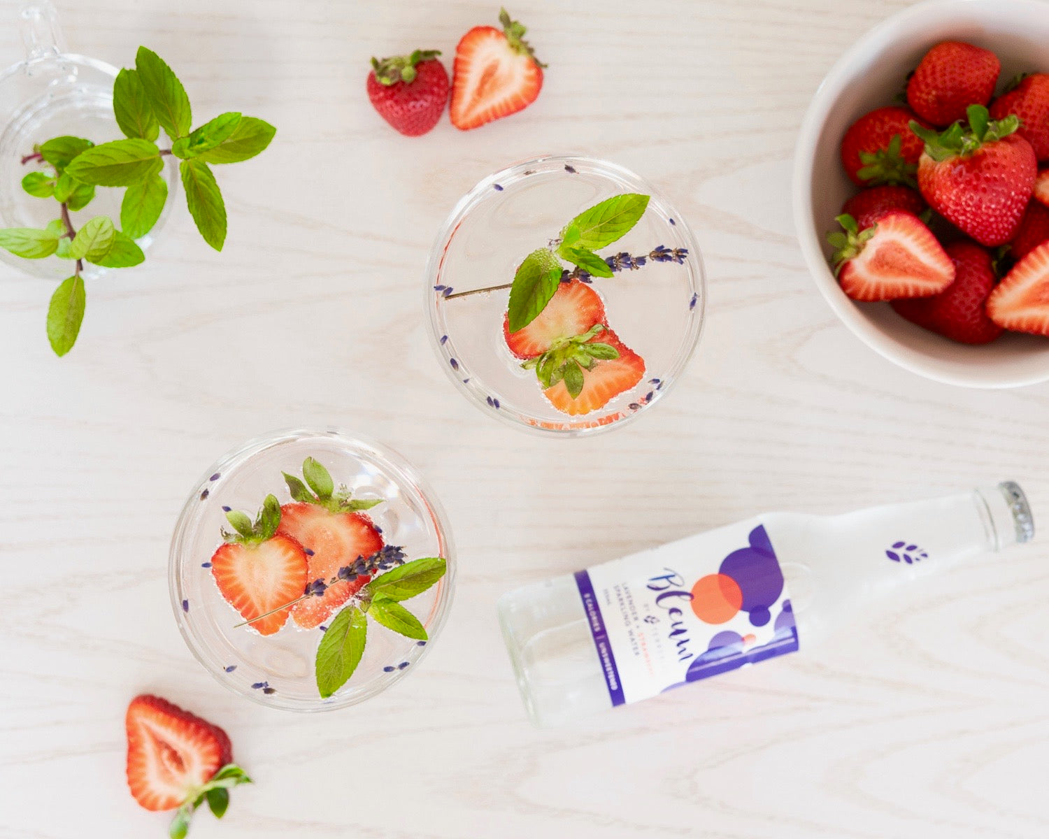 Glasses of sparkling water infused with lavender and strawberry on display on a white background with its bottle nearby. 