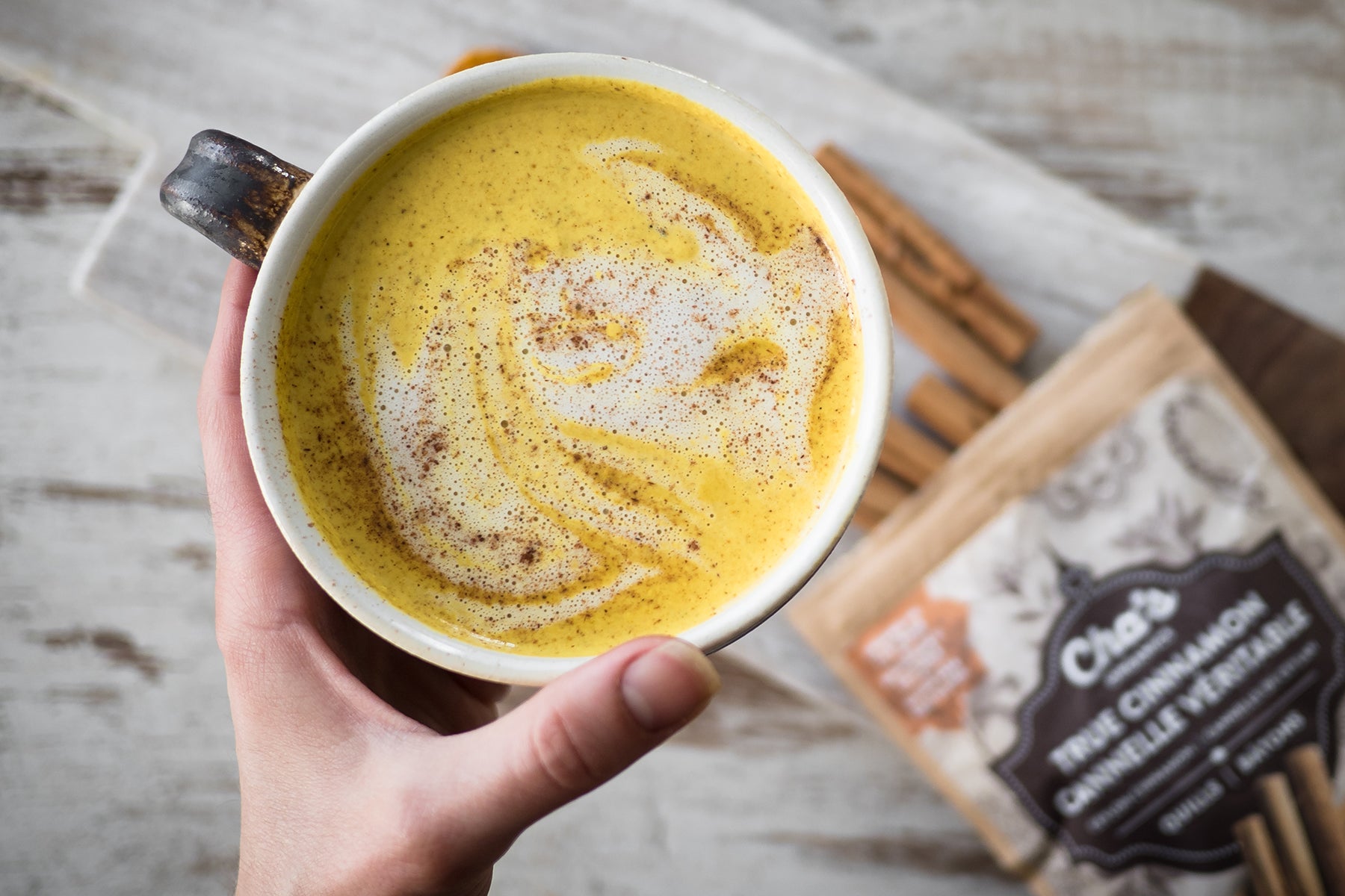 Aerial photograph of a caucasian woman's hand holding a cup of Cha's Organic's chai latte, over a wood surface. In the background, on the table beneath her hand is the branded Cha's Organics tea bag.