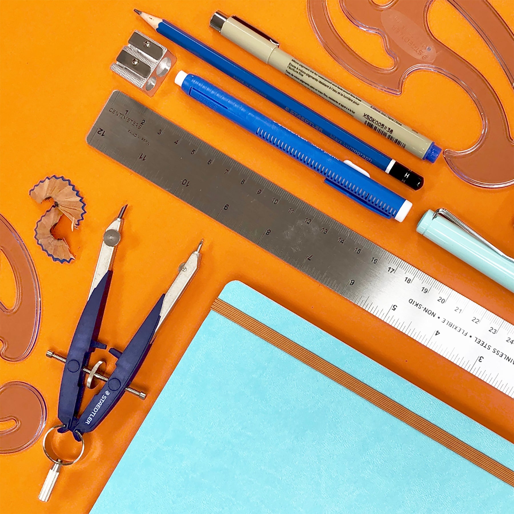 Aerial photograph of stationary supplies including a teal notebook, a ruler, pens and pencils and a compass, placed on top of a bright orange background.