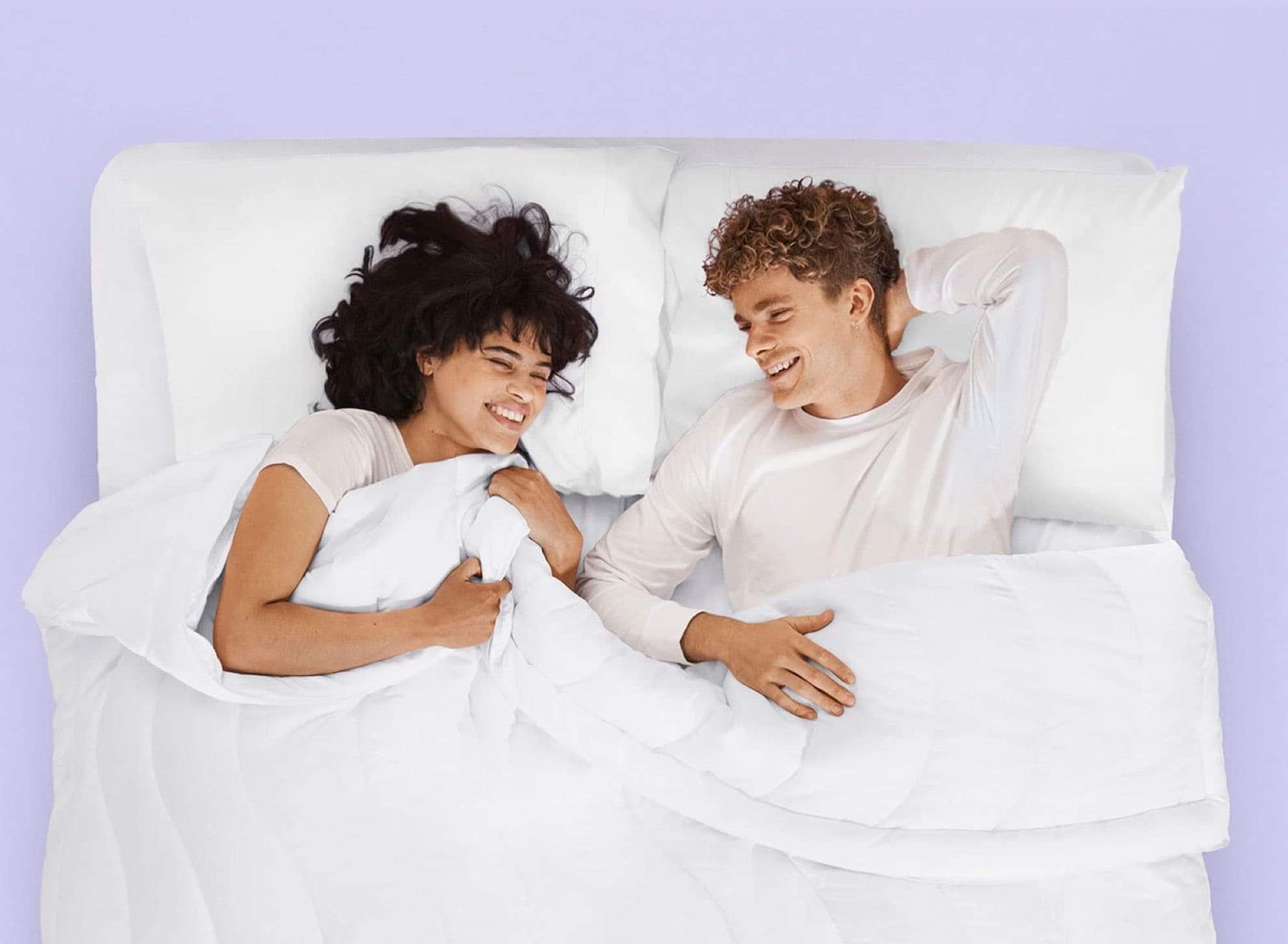 Two people lie in a white bed against a mauve background