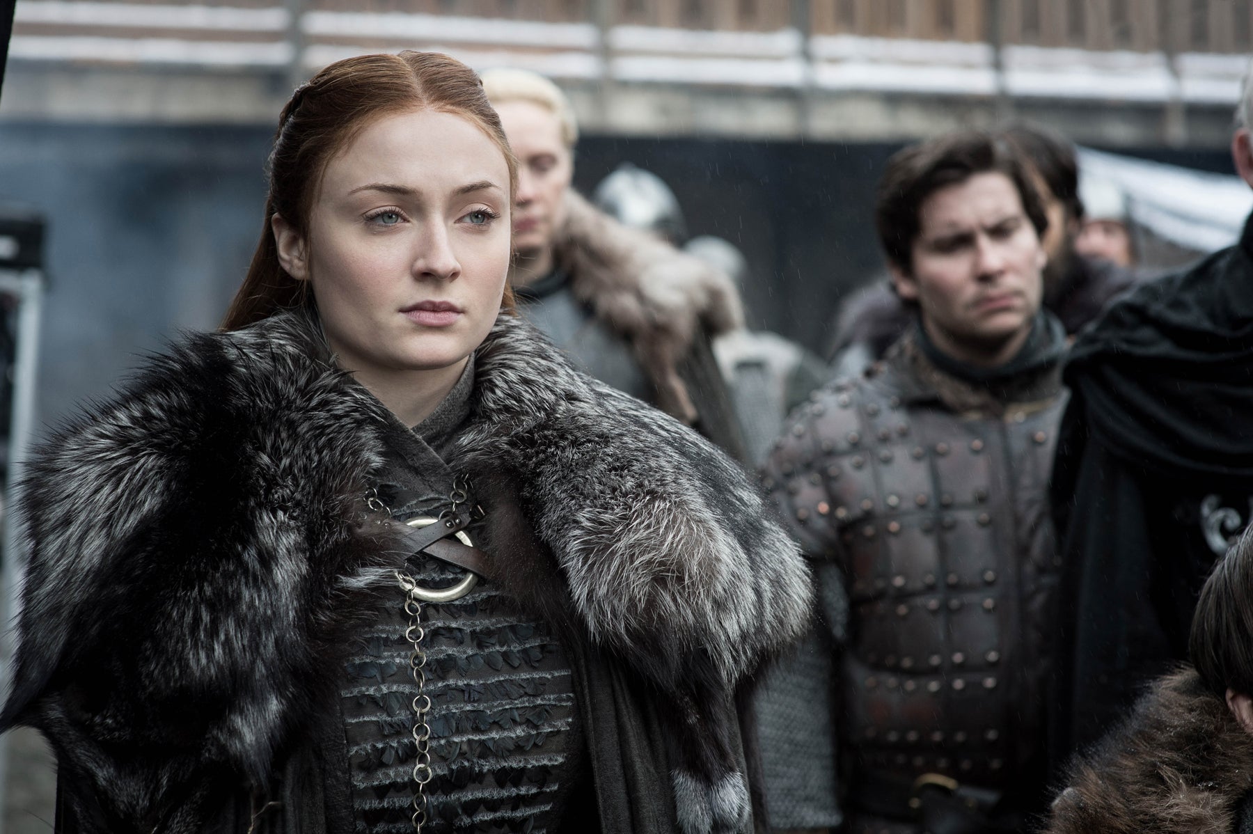 Sansa Stark stands in the courtyard of Winterfell, with Podrick and Brienne of Tarth in the background. Sansa looks off camera, glaring at someone approaching.