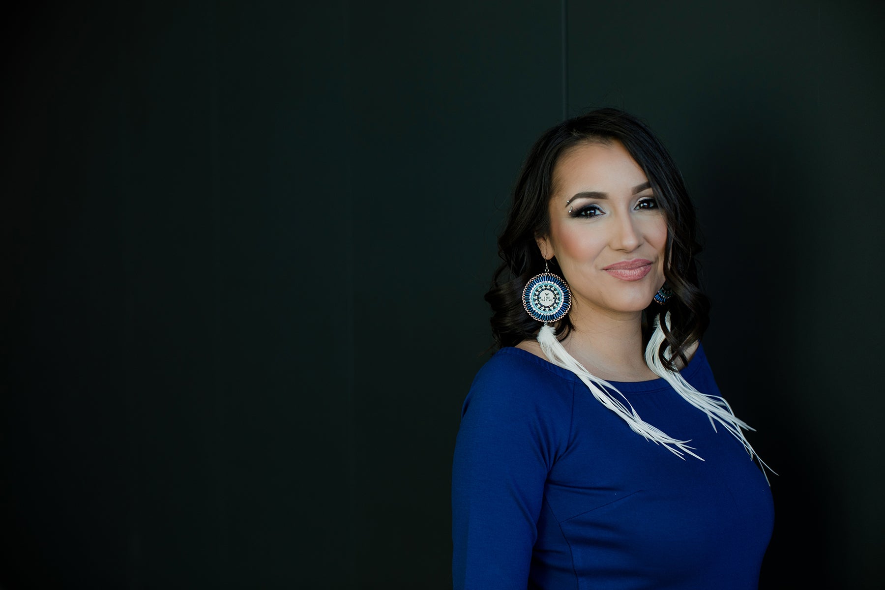 Portrait of designer Devon Fiddler of SheNative wearing blue and standing against a dark wall