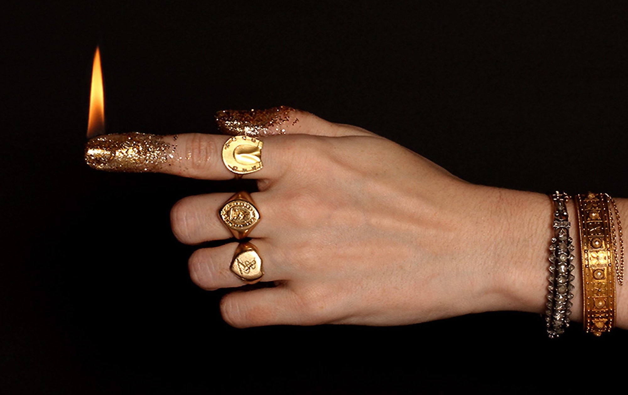 Hand wearing several vintage Erica Weiner rings. The index finger is coated in gold sparkles and is lit like a candle.