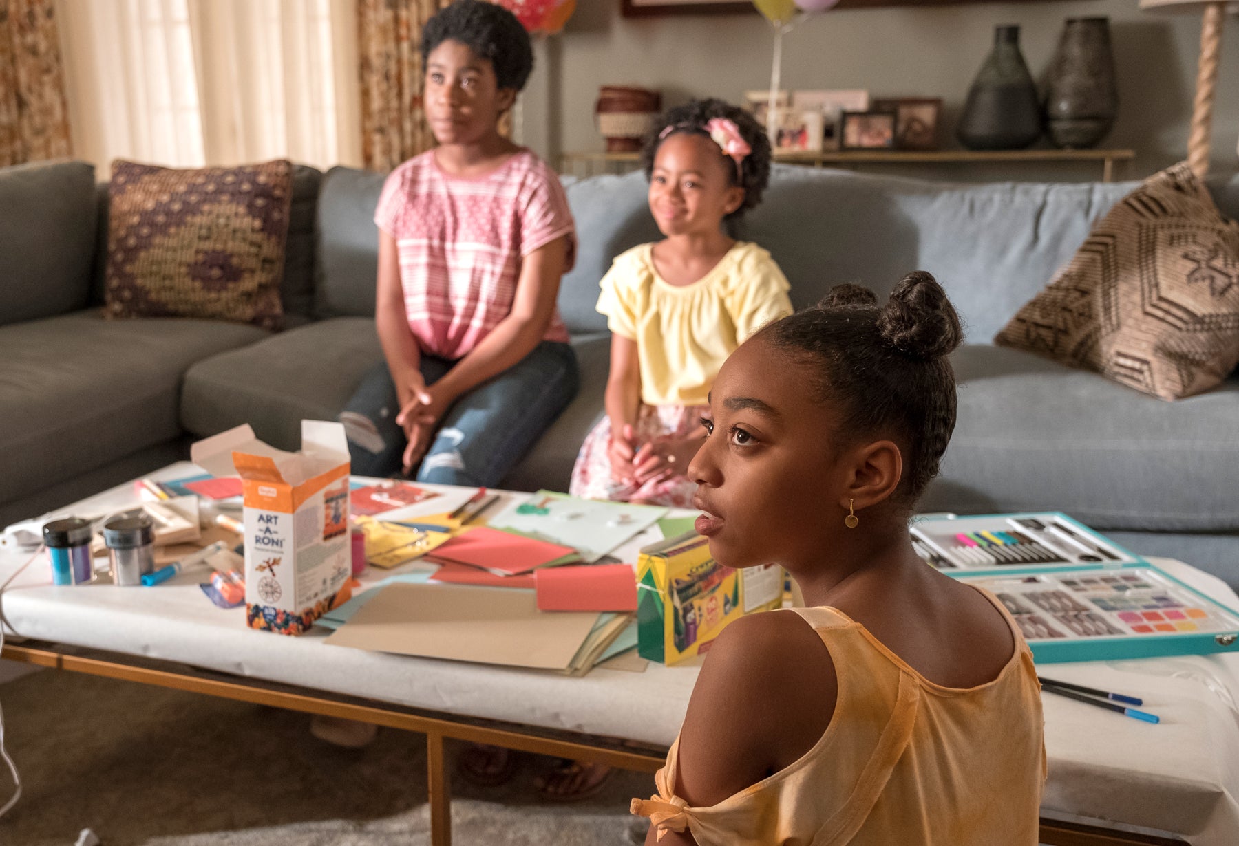 Deja (left), Annie Pearson (middle), and Tess Pearson (foreground) sit in a living room looking off camera in an episode of This Is Us. Crayons and craft supplies cover a coffee table between them.