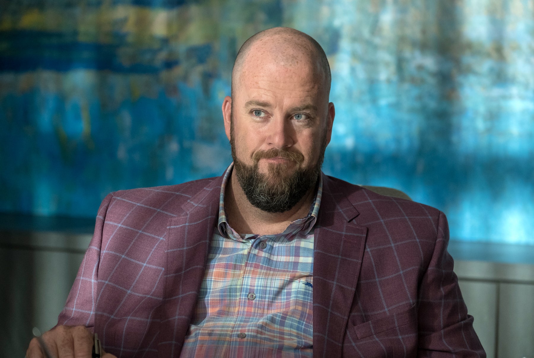 Toby (Chris Sullivan) sits in front of a blue mural wearing a plaid jacket and shirt in an episode of This Is Us.