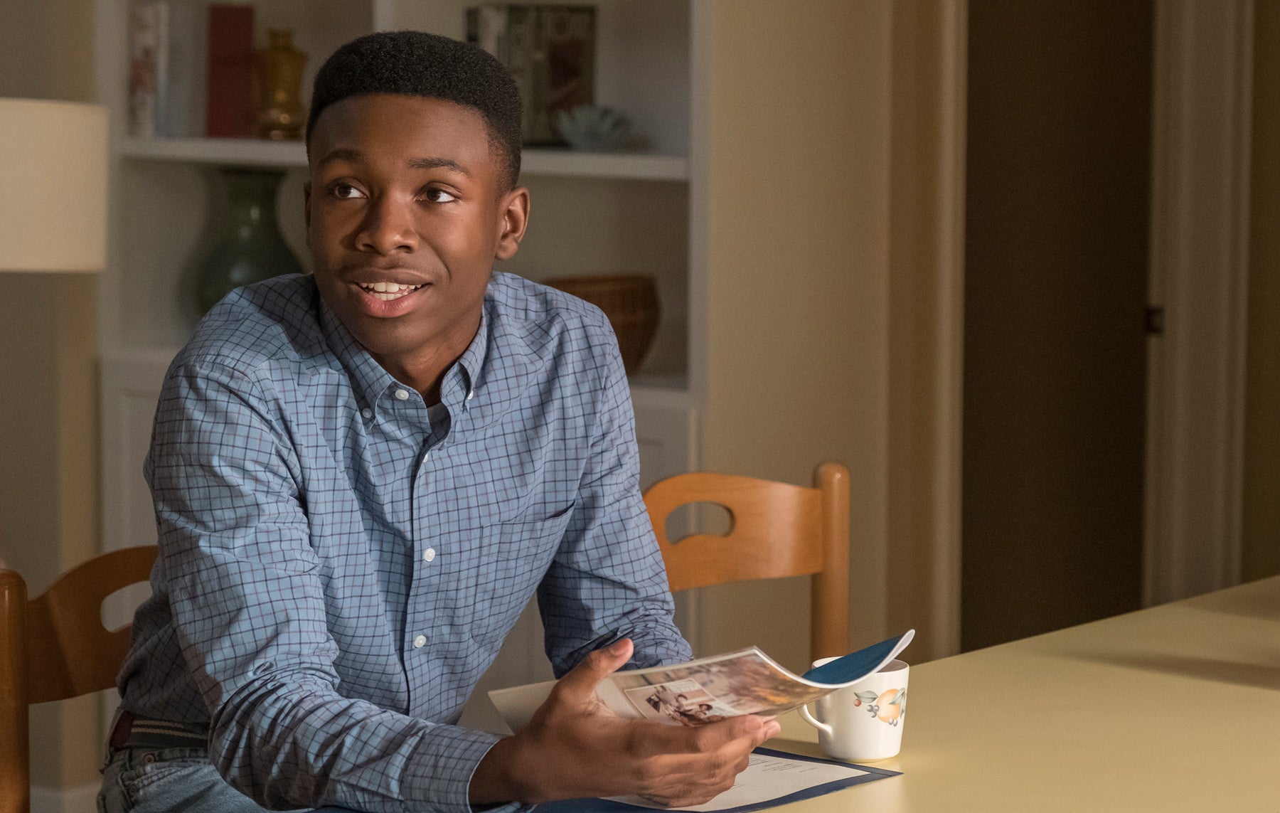 A young Randall Pearson (Niles Fitch) sits at a table with a coffee mug while holding a magazine in an episode of This Is Us.