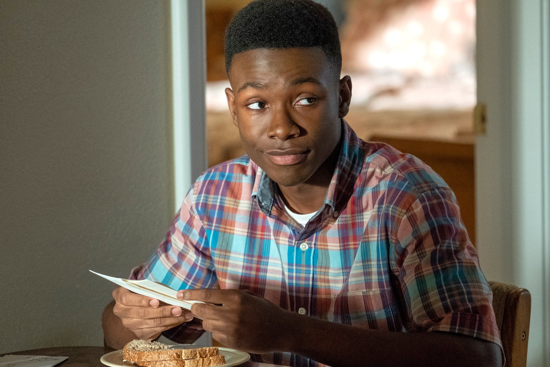 A young Randall Pearson (Niles Fitch) sits at a table with a sandwich while holding a paper in an episode of This Is Us.