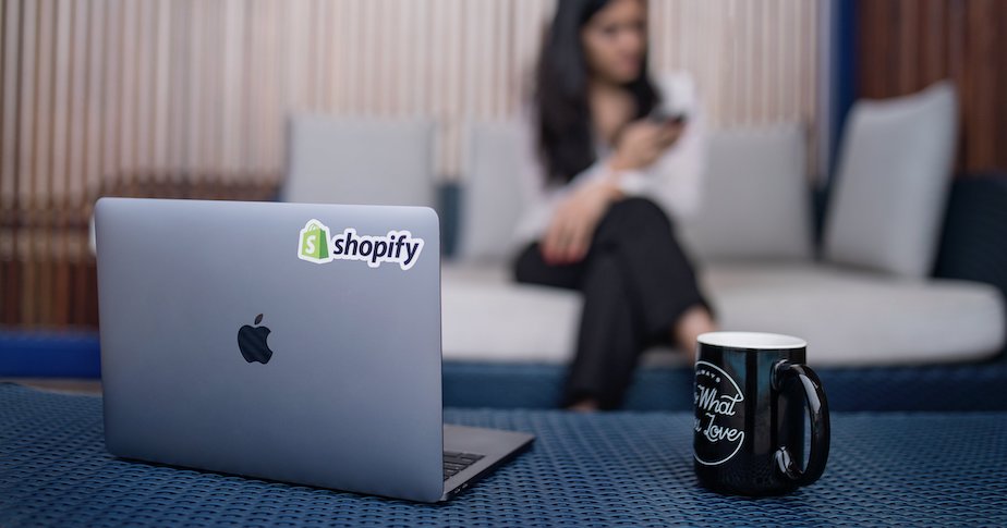 A laptop with a Shopify sticker backdropped by a women on a couch using a smartphone. 
