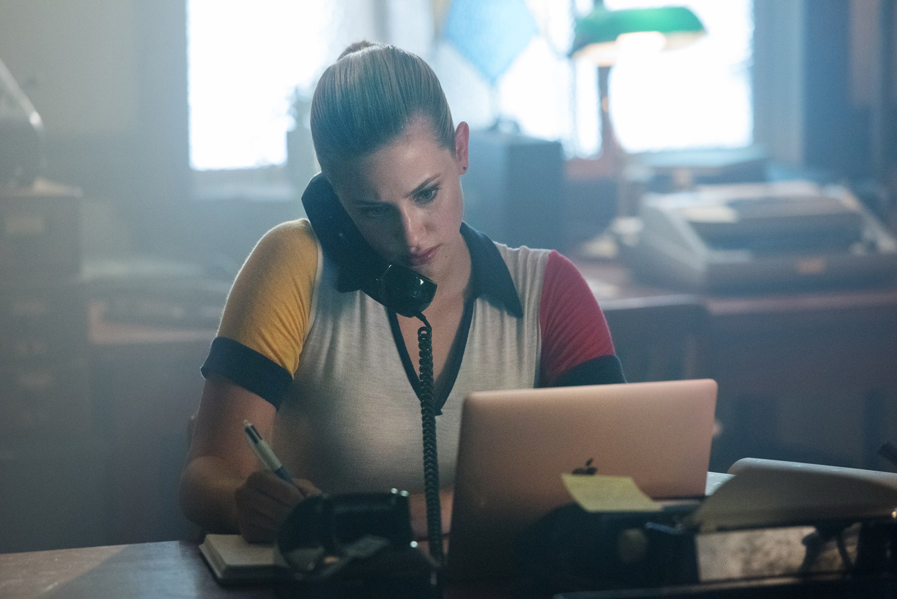 Betty Cooper talks on a phone and sits at a desk in front of a computer.