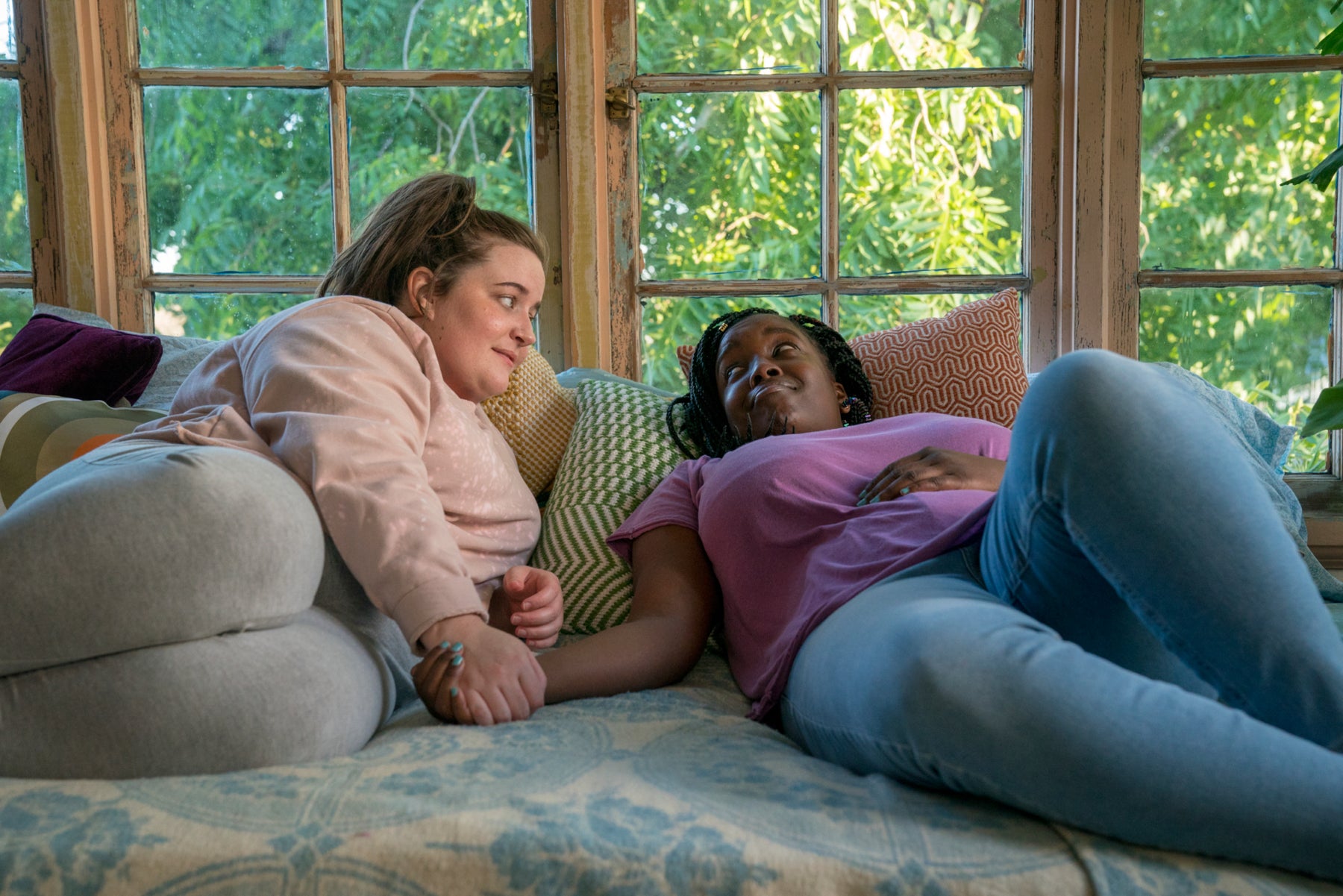 Annie and her roommate, Fran, lie on the bed, smiling at each other and holding hands. 