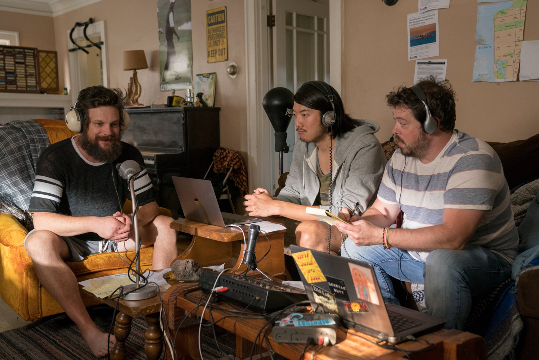 Ryan (Luka Jones) sits on the couch with headphones and in front of a microphone, taping a podcast with his two roommates. 