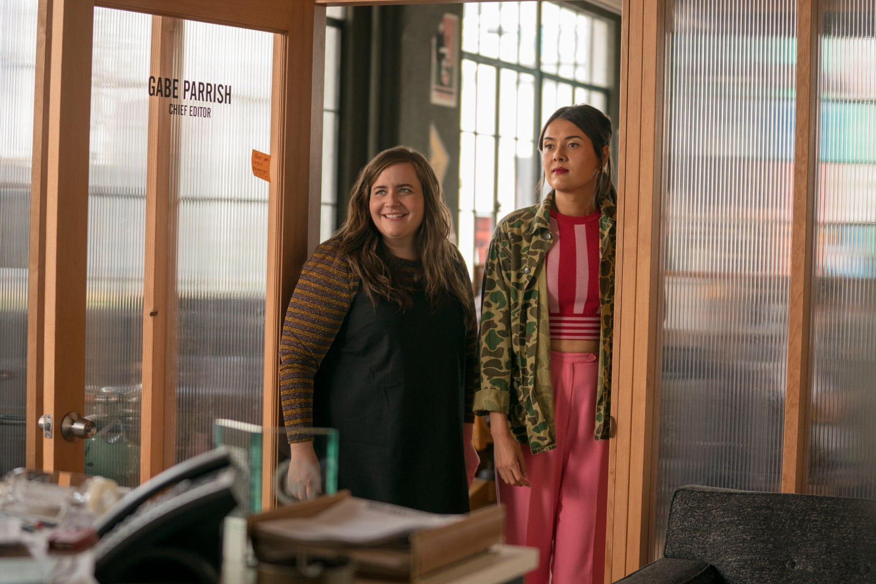 Annie stands in the doorway of her boss’s office, smiling, next to his assistant, Ruthi (Patti Harrison), who is not smiling. 