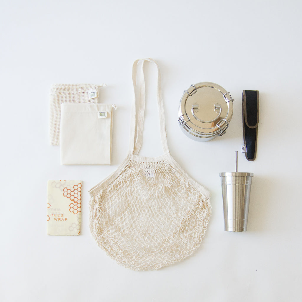 A zero-waste starter kit complete with French cotton net bag, cotton mesh produce bags, cotton muslin produce bags, beeswax wrappers, stainless steel lunchbox, stainless steel tumbler, and bamboo travel utensils. 
