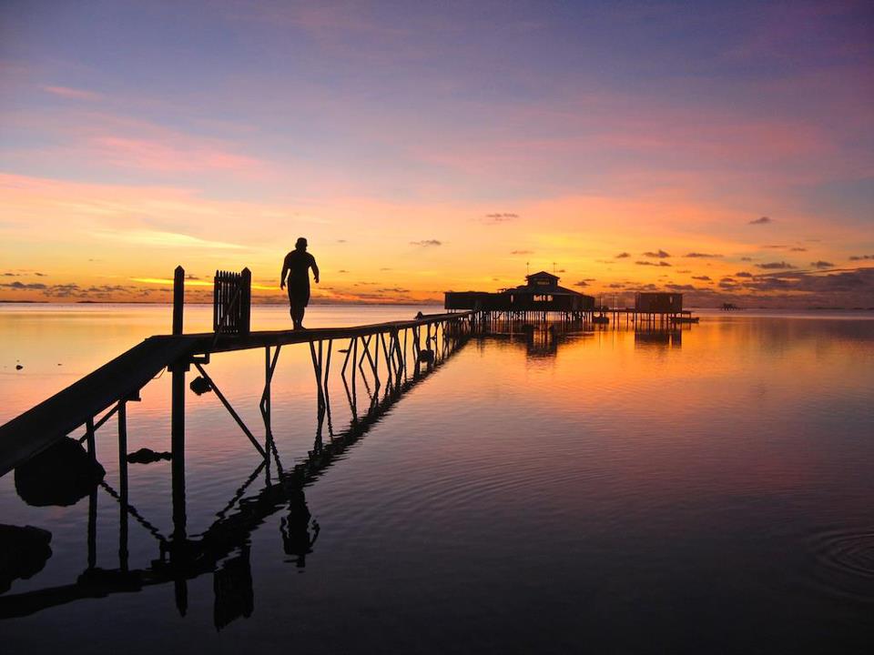 A photo of a man walking across a narrow bridge towards a building over the water. It is sunset and the calm lagoon reflects the orange and pink colors of the sky.