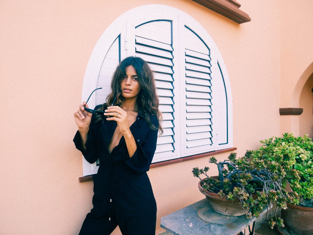 Sarah Qaisar holding sunglasses stood next to plant pots