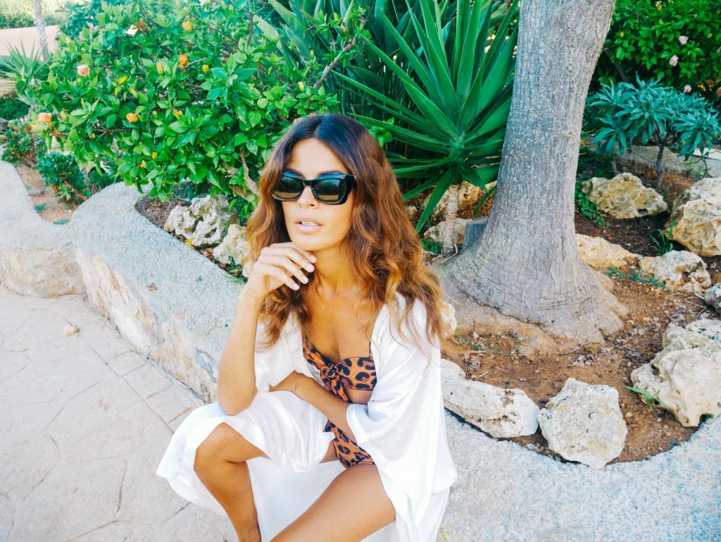 Sarah Qaisar sat on wall wearing sunglasses and leopard print bikini with white kaftan