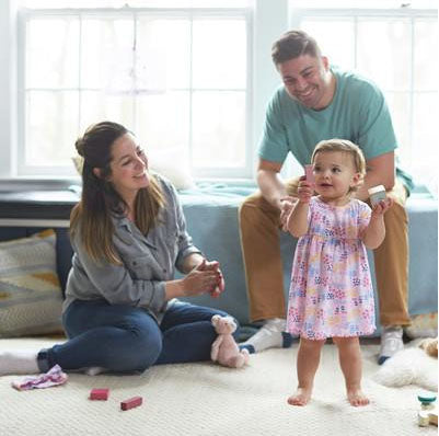 baby girl in dress stands with play blocks while mom and dad watch, smiling