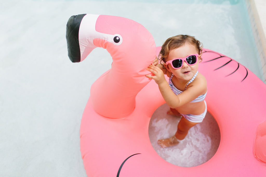 Toddler girl wearing Babiators sunglasses whiel floating in a pink flamingo pool toy