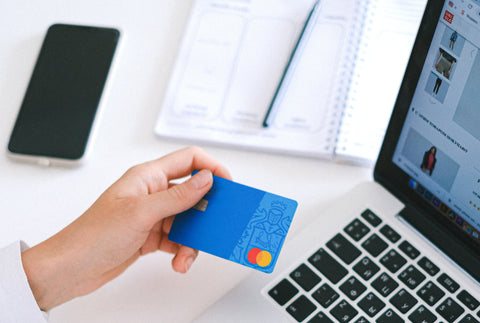 A close up of a hand holding a credit card. Also in the picture is a laptop, a notepad and pencil and a mobile phone