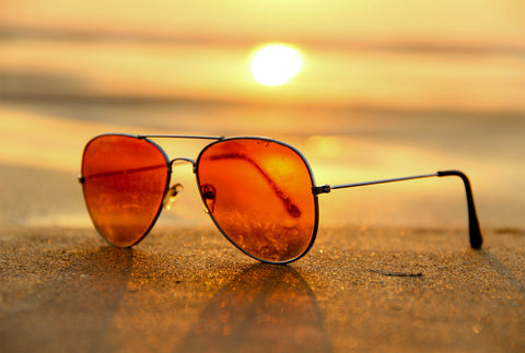 A pair orange lens sunglasses lying on some sand. The sun can be seen in the distance