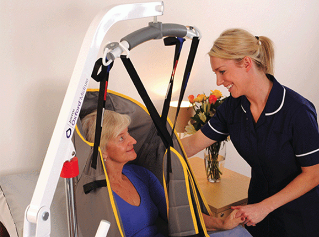 A woman in a sling beside a bed – she is being helped by a nurse