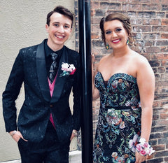 XO Babe and her date are standing next to a black pole. She has on a strapless black gown, with lots of beading and flowers on it.
