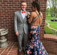 XO Babe Ashlynn is standing next to her date with her back facing us. She has a beautiful navy mermaid gown, with peach flowers on it. The back is open with criss-crossed straps. Her date has a grey tux on and they are standing in front of a brick wall.
