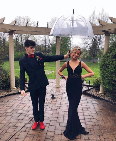 XO Babe, Chaste,  is standing under a clear umbrella, held by her date. She is in a black sheath gown with triangle cut outs at the wast. Her date is wearing an all black tux with bright red shoes and tie.