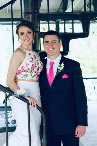 Olivia is standing next to her date. Scott, on a black spiral staircase She has on a white two piece gown. The top has pink, yellow, and green flowers embroidered on it. Scott is wearing a black tux, with a hot pink tie.