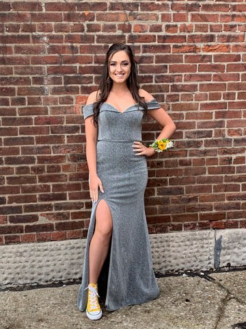 XO Babe, Mackenzie, is standing in front of a brick wall. She has a silver sheath gown on and bright yellow Converse high top shoes on. She also is wearing a corsage with yellow sunflowers in it.