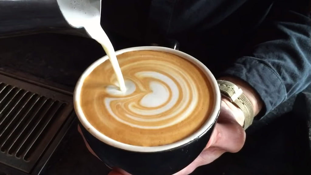 Milk pouring from a stainless steel frothing pitcher into a latte to make latte art into a cappuccino cup.