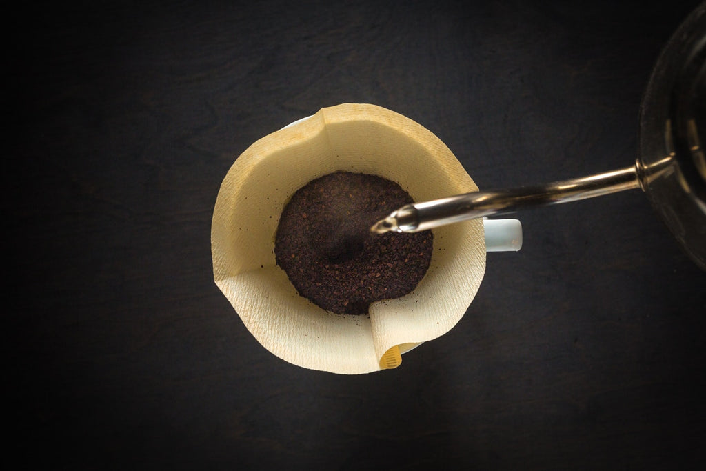 A bird's eye view of the neck of a silver kettle pouring water into a wet coffee filter with ground coffee inside of it, the handle of a white mug can be seen below the filter, too.