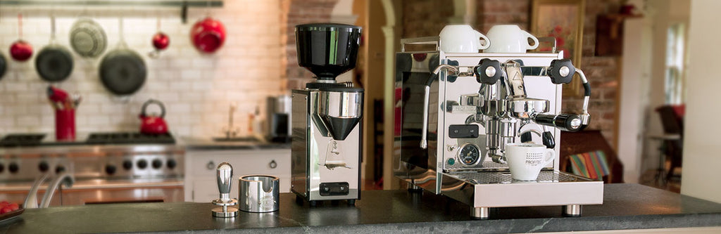 A lifestyle image of a Profitec Pro 600 and a Profitec Pro T64 Grinder on a top of a counter, with an ECM pressurized tamper and ECM Tamping station next to it. There is a kitchen in the background with pots and pans hanging from the walls. 