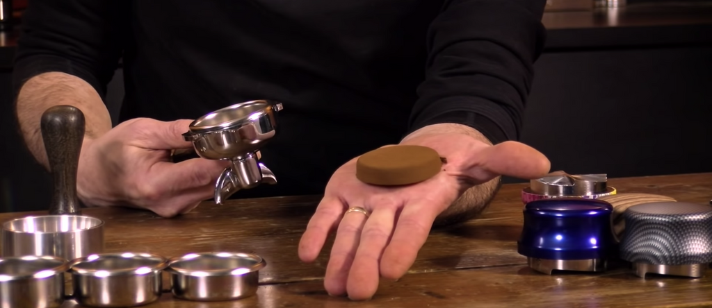 A dry puck of coffee in an open palm, with an empty portafilter in the other hand. There is a wooden table underneath the hand with three Asso Jacks one one side, and empty filter baskets and other accessories on the other side.  