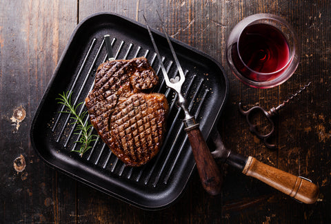 Ribeye Steak with Rosemary in a pan. Surrounded by a glass of red whine and a grilling fork.