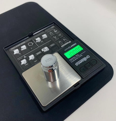The Truweigh Mini Classic digital scale sits on a black mat on a white countertop with a 100g calibration weight on the platform. The green screen reads 100.00g