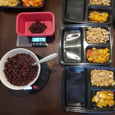 A meal prep station is shown from above. A bowl of beans is being measured into the Truweigh Crimson weighing bowl on the scale, and four meal prep containers are set up and already filled with protein and vegetables.